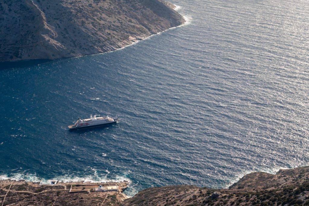 Aerial view of a large ship near a rugged coastline with blue waters, showing a remote landscape with sparse infrastructure under bright sunlight.