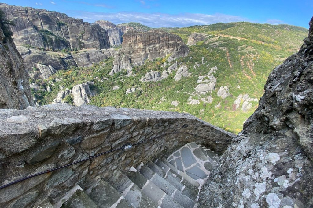 An ancient stone stairway descends a steep cliffside, offering a breathtaking view of layered mountains and lush valleys under a clear blue sky.