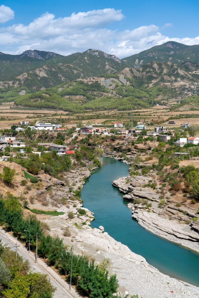 A serene river flows through a rocky landscape, passing a small village with houses scattered between roads, fields, and gently rolling hills.
