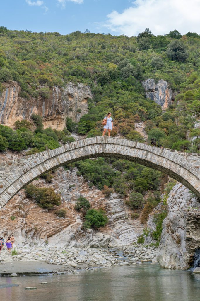 An old arched stone bridge spans a river with a person standing on top, surrounded by a rugged, tree-covered landscape and blue skies above.