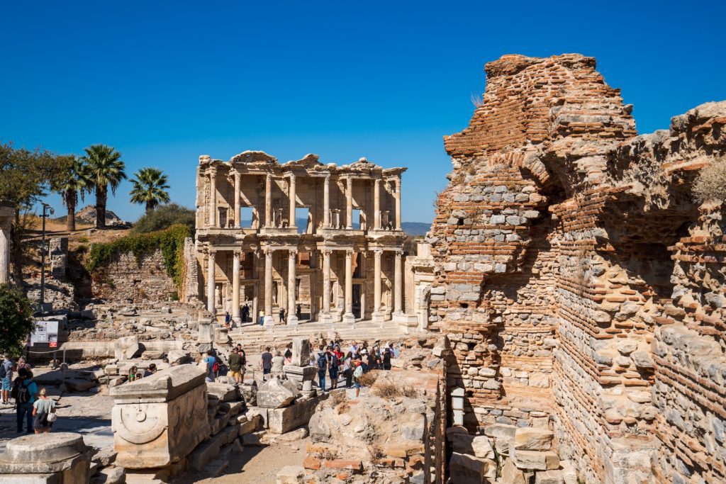This image shows the ancient Library of Celsus in Ephesus, Turkey, with visitors exploring ruins and palm trees in the background under a clear blue sky.