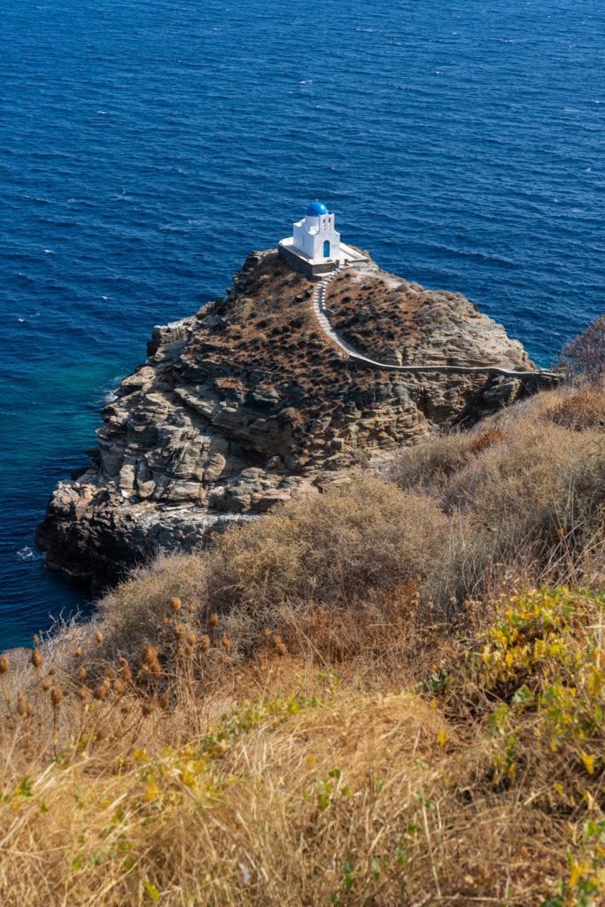 A small white chapel with a blue dome sits atop a rocky promontory surrounded by the blue sea, connected by a winding pathway with dry vegetation.