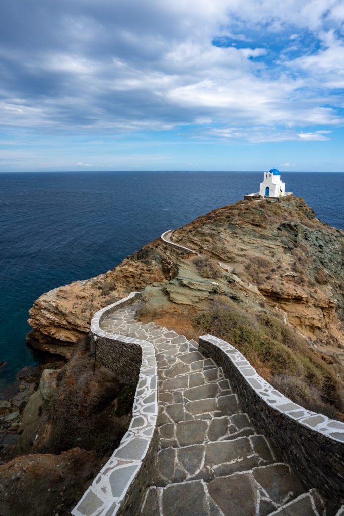 A winding stone path leads to a small white chapel perched on a rocky cliff overlooking the expansive blue sea under a cloudy sky.