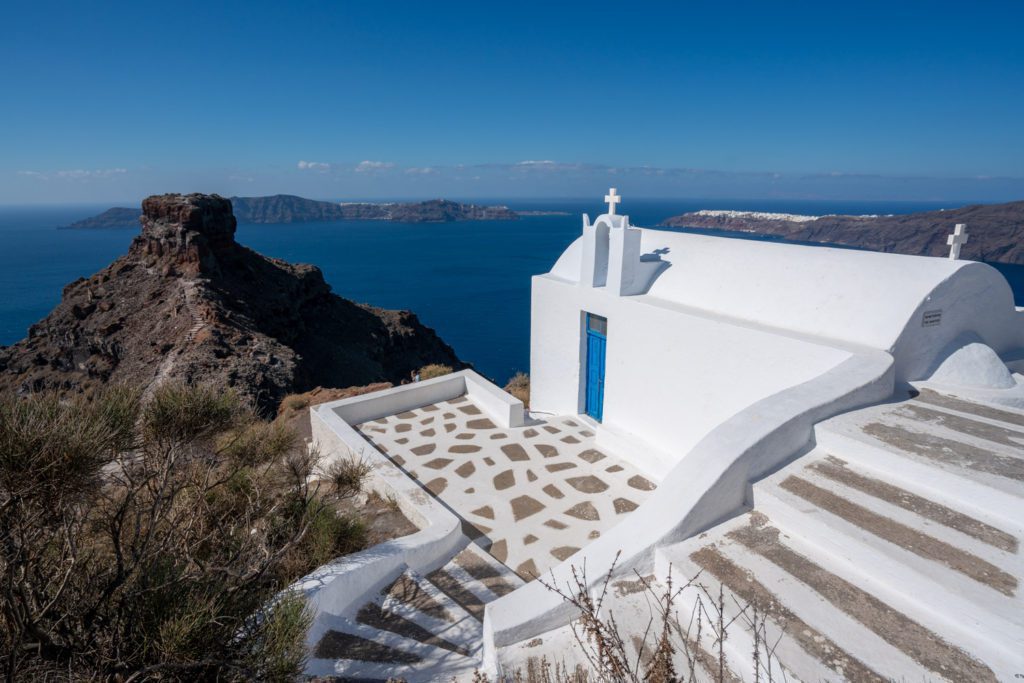 A small white church with a blue door overlooks the sea, nestled on a hill above the rugged coastline. Steps lead up to a decorated patio.