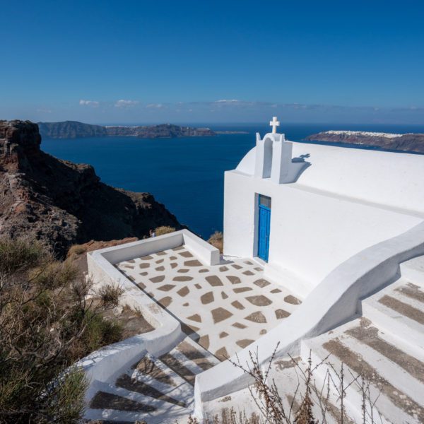A small white church with a blue door overlooks the sea, nestled on a hill above the rugged coastline. Steps lead up to a decorated patio.
