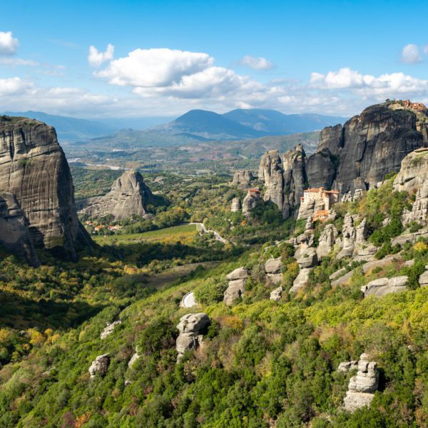 This image shows a stunning landscape with towering rock formations and a monastery perched atop a cliff amidst a lush, green forested area, under a partly cloudy sky.