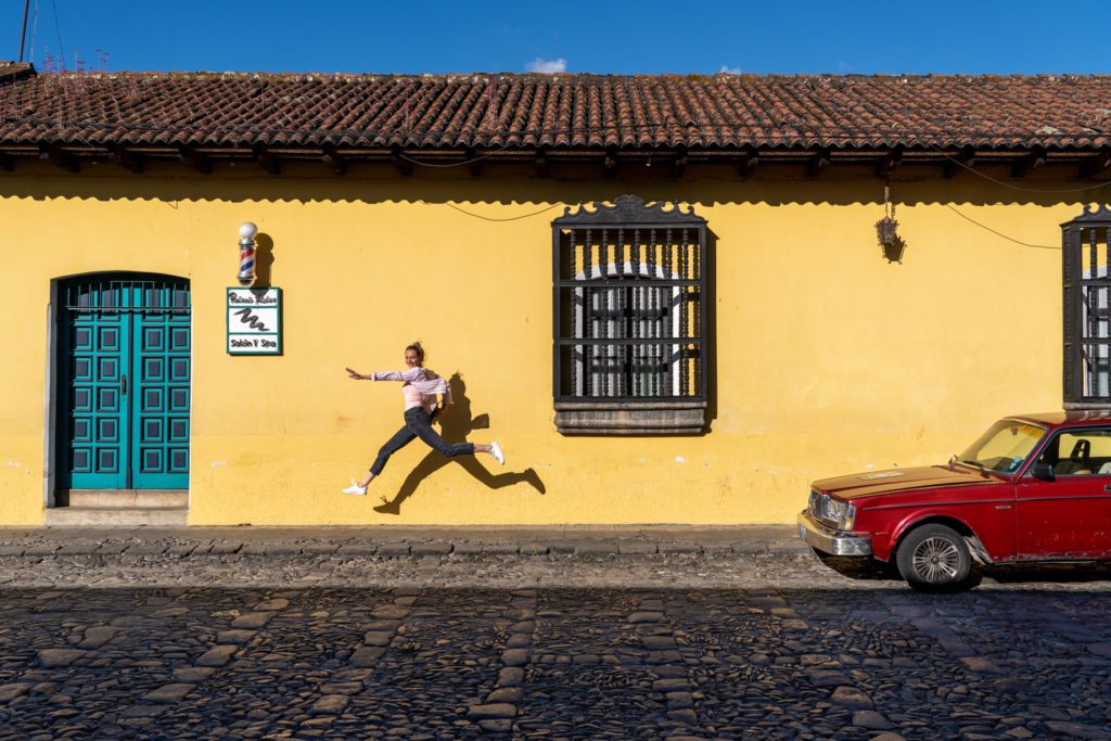 A person is mid-jump on a cobblestone street beside a yellow wall, near a classic red car, under a bright blue sky.