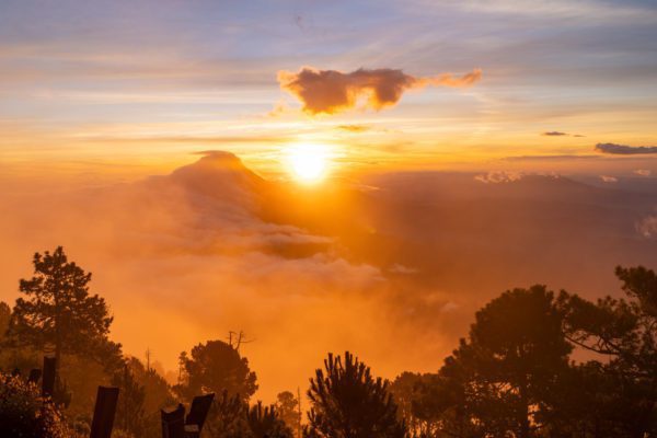 A breathtaking sunrise with golden hues breaking through a dense, cloud-filled sky above a silhouette of a mountain range and forested area in the foreground.