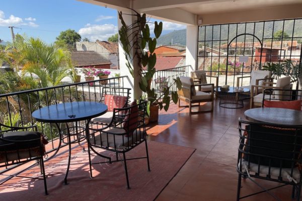 This image shows a sunny, outdoor terrace with wrought iron furniture, potted plants, a tile floor, protective railing, and a view of a town under a blue sky.