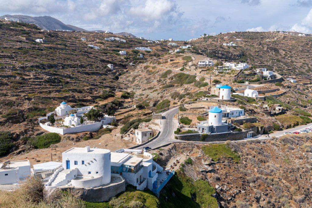 A winding road traverses a hilly landscape dotted with white buildings, some with blue domes, under a partial cloud cover on a sunny day.