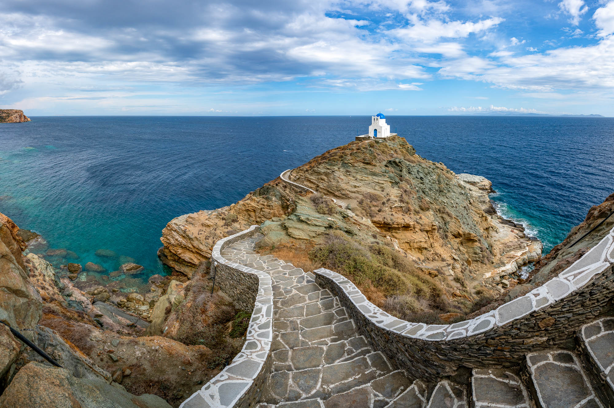 A winding stone pathway leads to a small white lighthouse perched on a rugged cliff overlooking the serene blue sea under a partly cloudy sky.