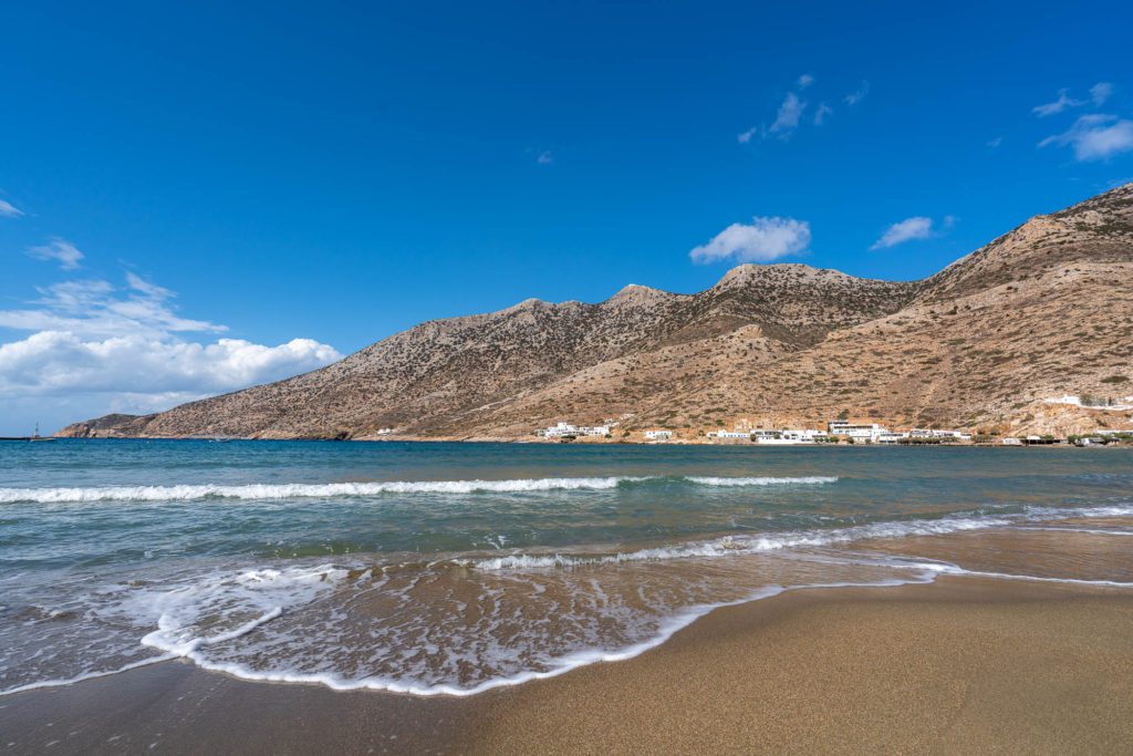 This is a serene beach scene showing gentle waves lapping onto sandy shore, with a backdrop of rugged mountains under a partly cloudy sky.