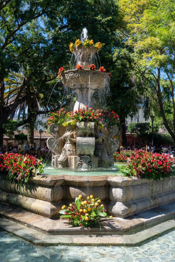 An ornate stone fountain, adorned with fresh flowers, cascades water amidst lush greenery under a bright, sunny sky in a serene park setting.