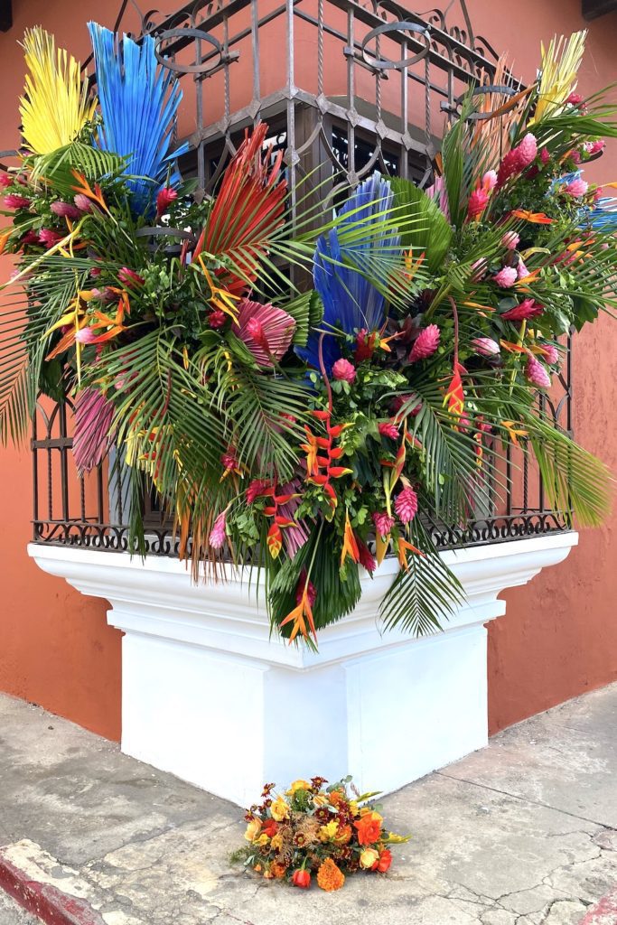 A vibrant floral arrangement bursts with color around a window grille, atop a white pedestal. Below, a smaller bouquet complements the display against peach walls.