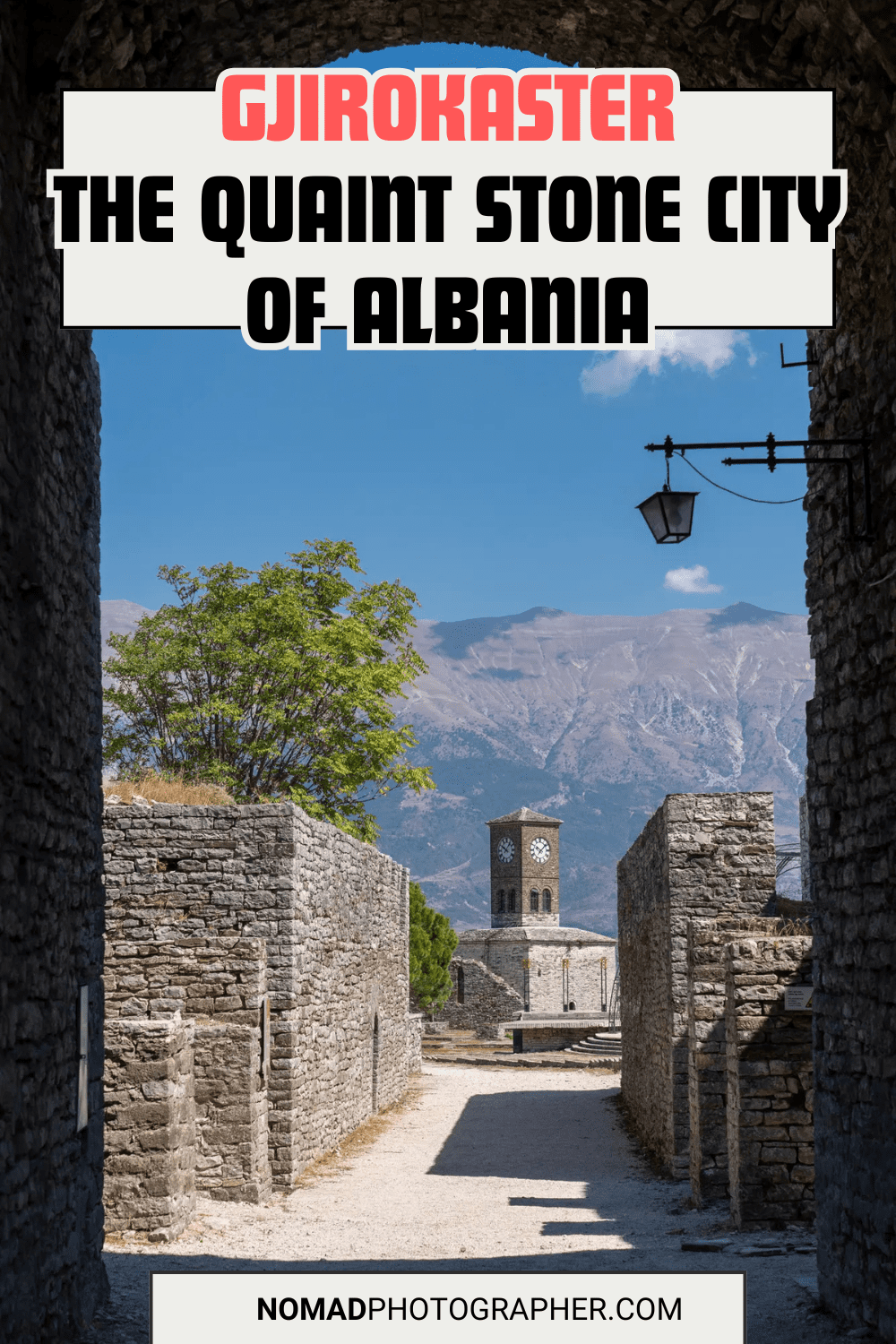 Historic stone buildings in Gjirokastër, Albania, with a clock tower in the background and mountainous landscape. A path leads through weathered stone walls.