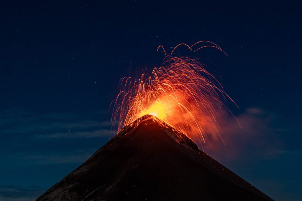 An active volcano at night, erupting with vibrant orange lava and emitting trails of glowing sparks against a dark blue sky studded with stars.