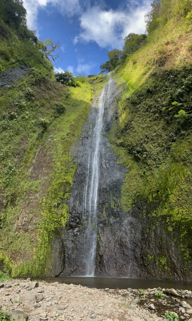 A tall, slender waterfall cascades down a mossy cliff into a small pool below, surrounded by lush vegetation under a blue sky with scattered clouds.