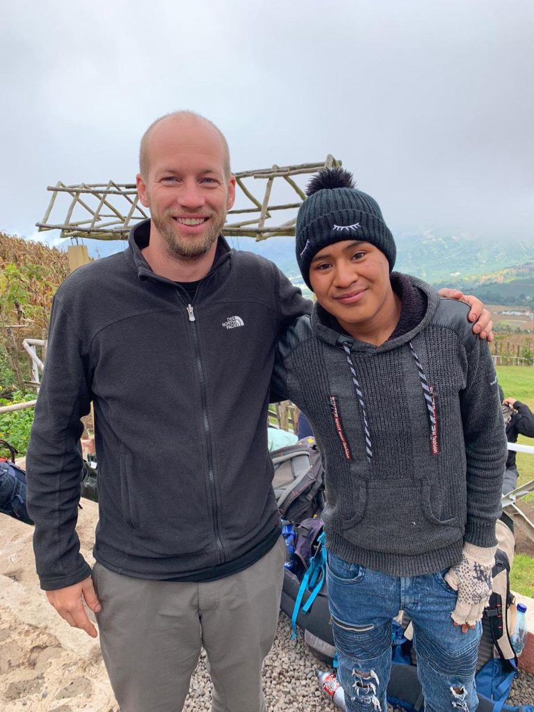 Two people are smiling for the camera in an outdoor setting. Both are wearing casual, cool-weather clothing, with a natural landscape in the background.