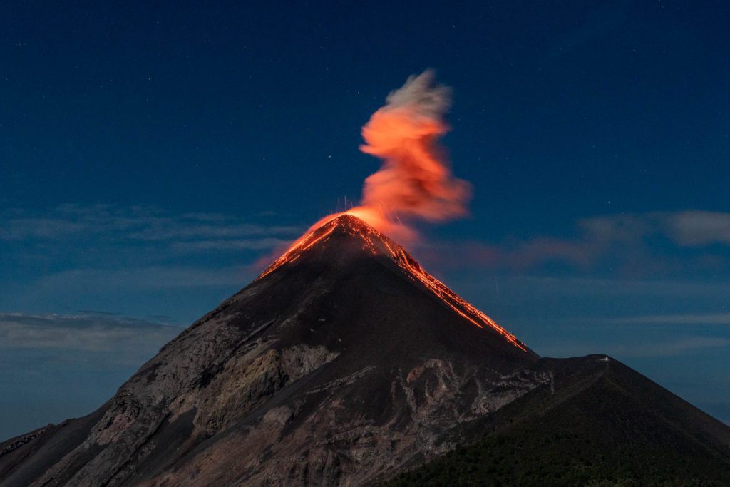 A volcano is erupting at twilight, with glowing lava streaming down and a fiery plume against the darkening sky. The scene shows nature's powerful forces.