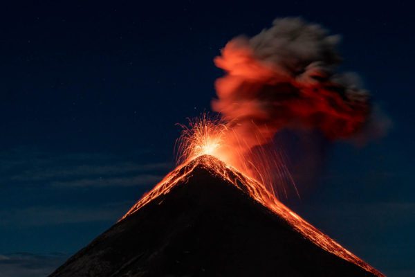 A volcano erupts at night, spewing lava and ash into the sky, with a glowing red flow down its slopes against a dark blue backdrop.