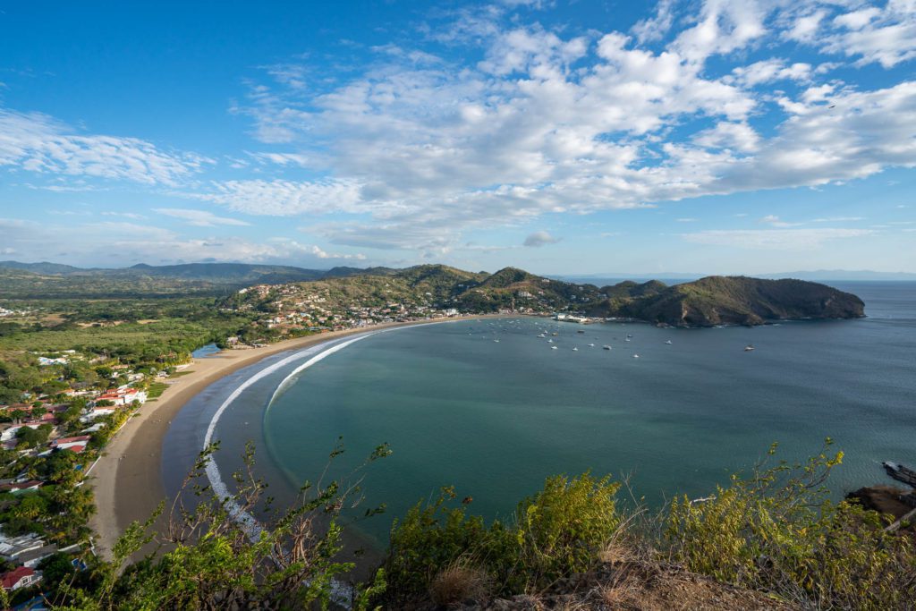 A scenic coastal landscape with a curved beachfront, calm bay with boats, lush greenery, a small town, and hills under a partly cloudy sky.