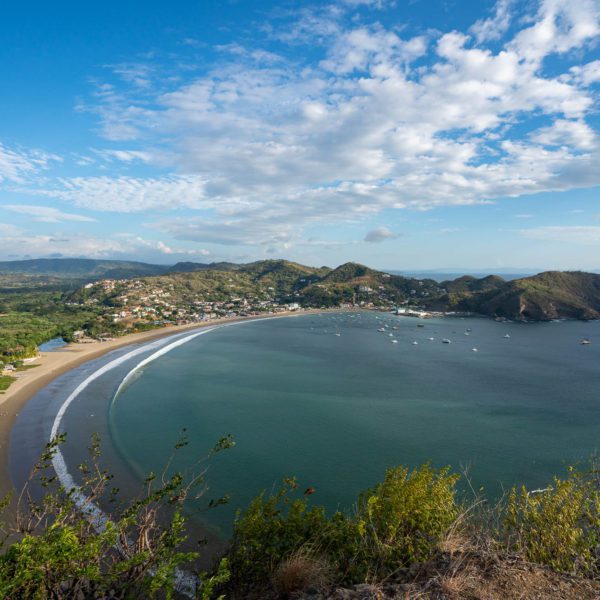 A scenic coastal landscape with a curved beachfront, calm bay with boats, lush greenery, a small town, and hills under a partly cloudy sky.