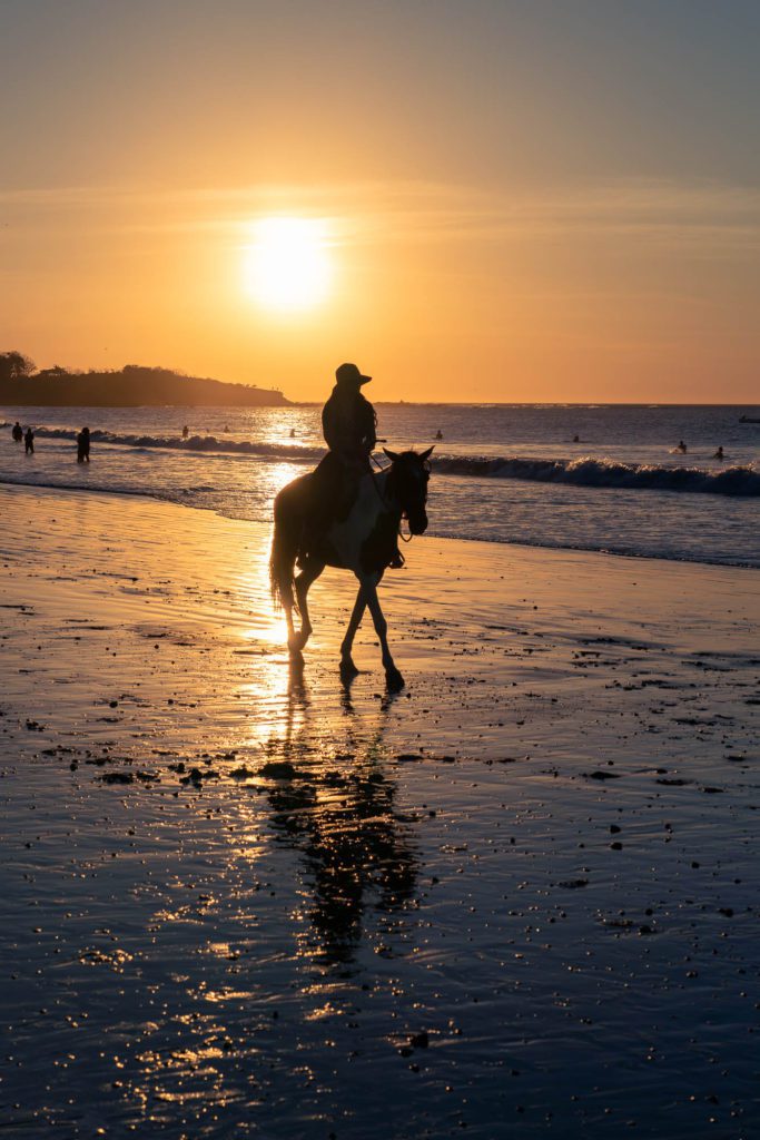 A person rides horseback along a shimmering beach at sunset, with the sun casting a warm glow and silhouettes of other beachgoers visible in the distance.