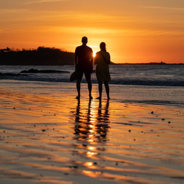 Two people stand on a wet beach at sunset, silhouetted against the orange sky, holding hands and gazing out towards the sea horizon.