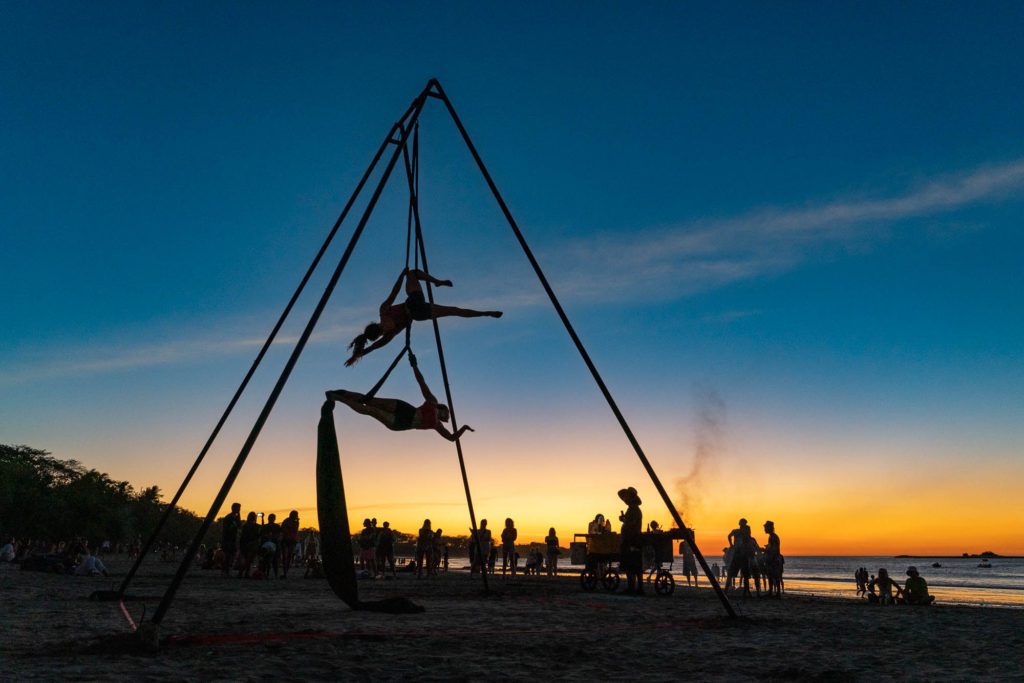 This image shows an aerial silk performer suspended from a tripod frame on a beach at dusk, silhouetted against a colorful sunset, with onlookers nearby.