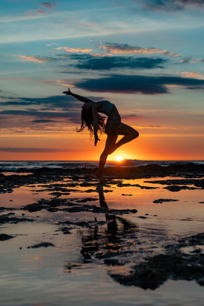 A person performs a yoga pose on a rocky beach against a colorful sunset sky, with the sun's reflection in the water.