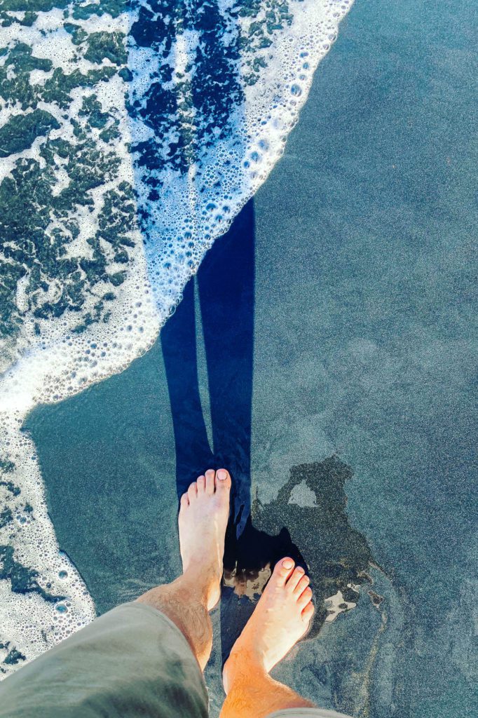 A person's bare feet at the edge of the surf on a sandy beach; the oncoming waves create a white foam; sunny day with shadows.