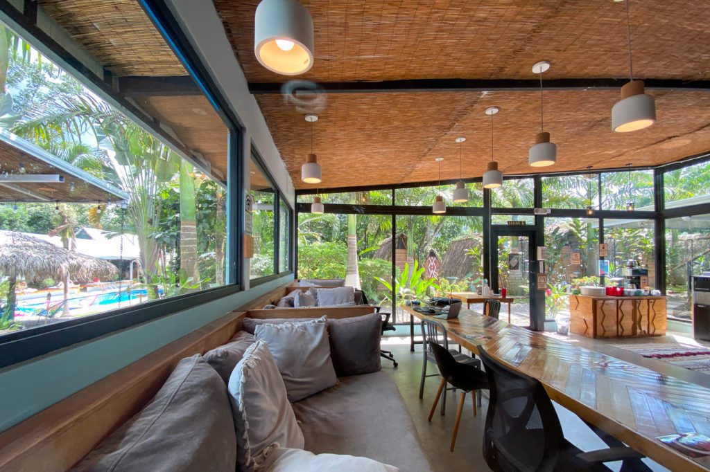 This is a modern, tropical interior with a long wooden table, comfortable seating, and pendant lights, surrounded by large windows revealing lush greenery outside.