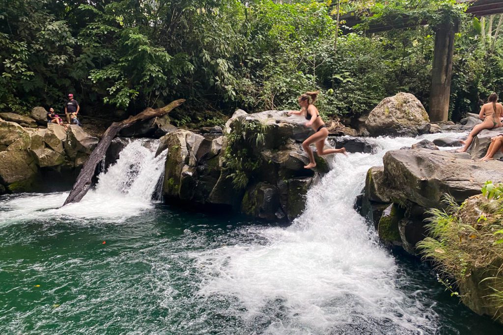 A person is mid-jump into a clear, forest-surrounded river with small waterfalls. Other individuals relax on the rocks, and lush greenery abounds.