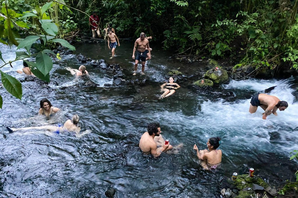 A group of people is enjoying a day at a natural stream surrounded by lush greenery. Some are swimming, while others are jumping or conversing.