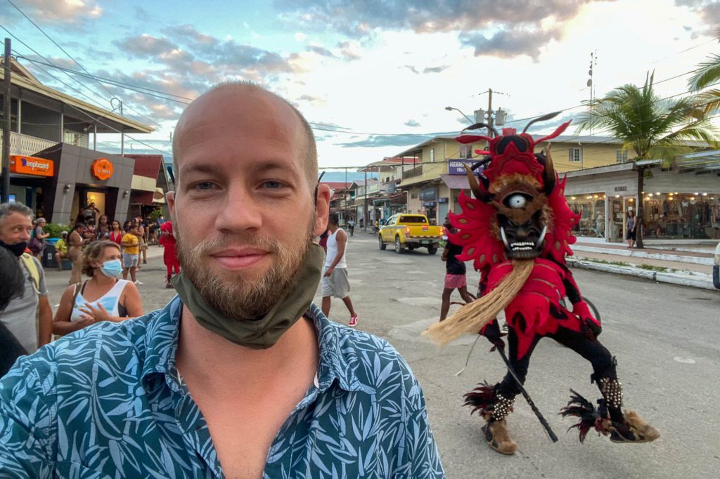 A person is taking a selfie on a lively street; behind them, a figure in a red, demonic costume with a mask parades by. The setting sun casts a warm light.