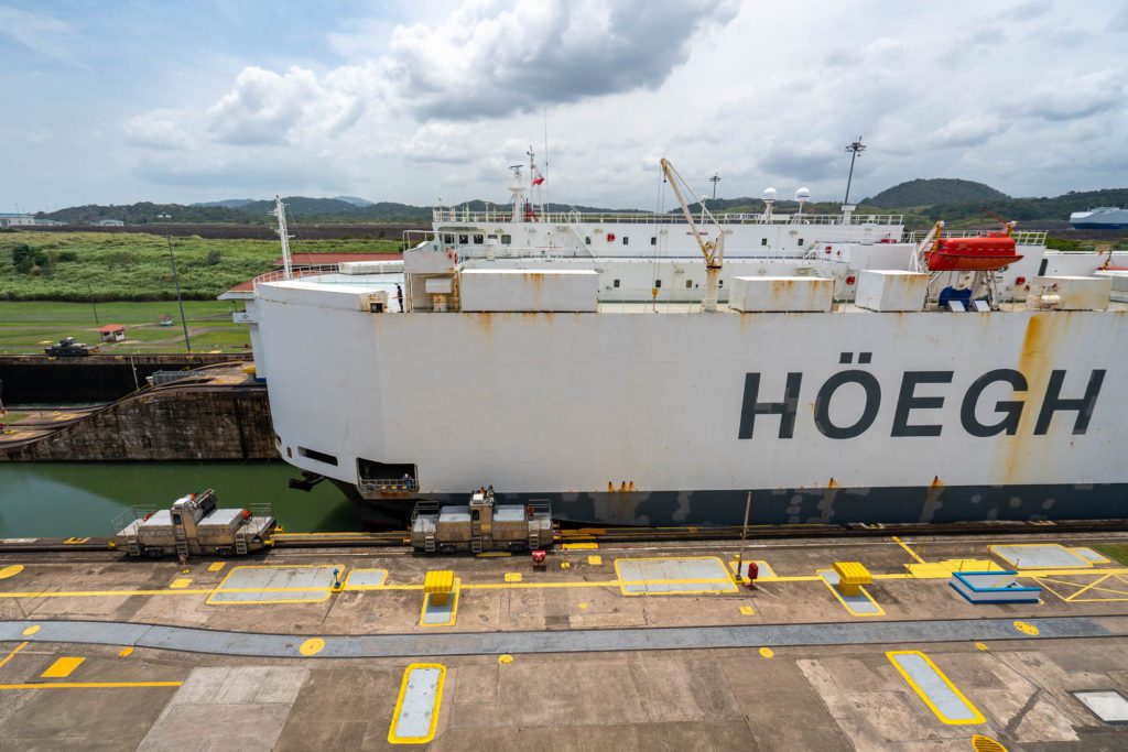 A large white cargo ship with "HÖEGH" written on the side is docked in a lock, with green hills and a cloudy sky in the background.