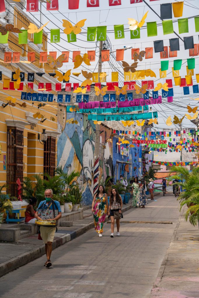 A vibrant street scene with colorful banners, people walking, and bright building facades. Artistic murals add to the lively atmosphere of the locality.