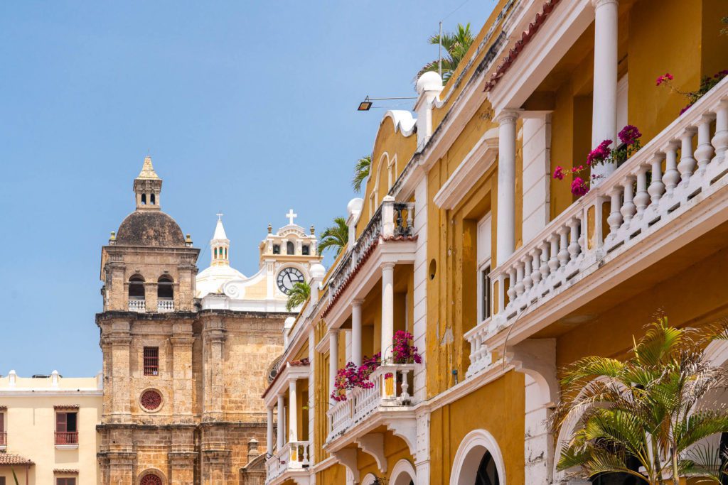 A picturesque scene with colonial architecture featuring yellow buildings, balconies with pink flowers, and a historic church with domes in the background under a blue sky.