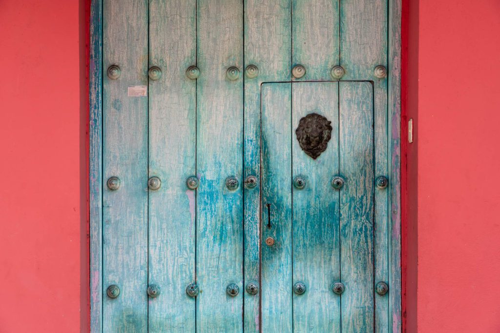 This image displays a weathered blue wooden door with decorative ironwork and metal studs, set in a vibrant pink wall. The door shows signs of aging.