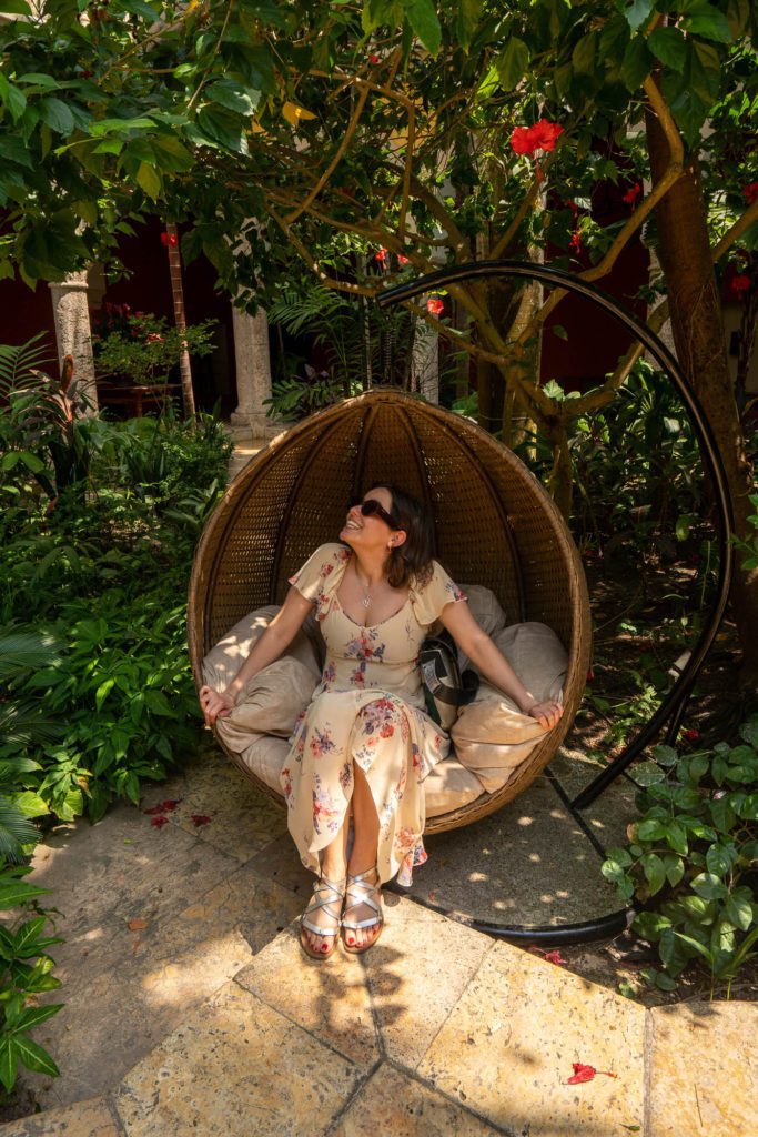A person relaxes in a hanging egg chair, surrounded by lush greenery and hibiscus flowers, sunlight filtering through the leaves overhead.