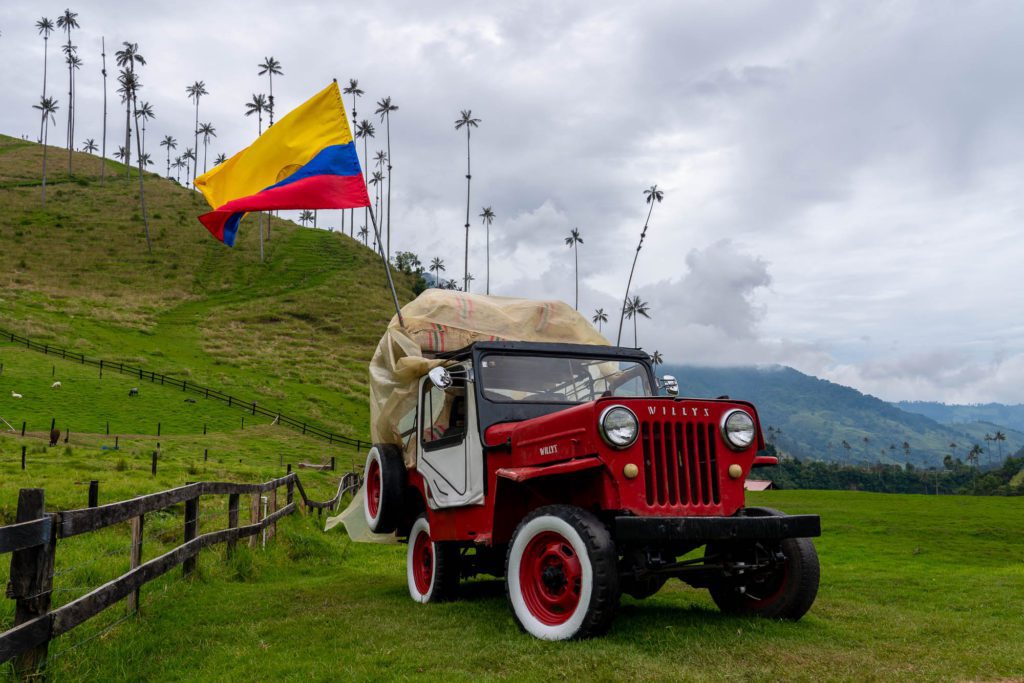 An old red and white jeep is loaded with goods, parked in lush green terrain under a cloudy sky, near a large Colombian flag waving atop a pole.
