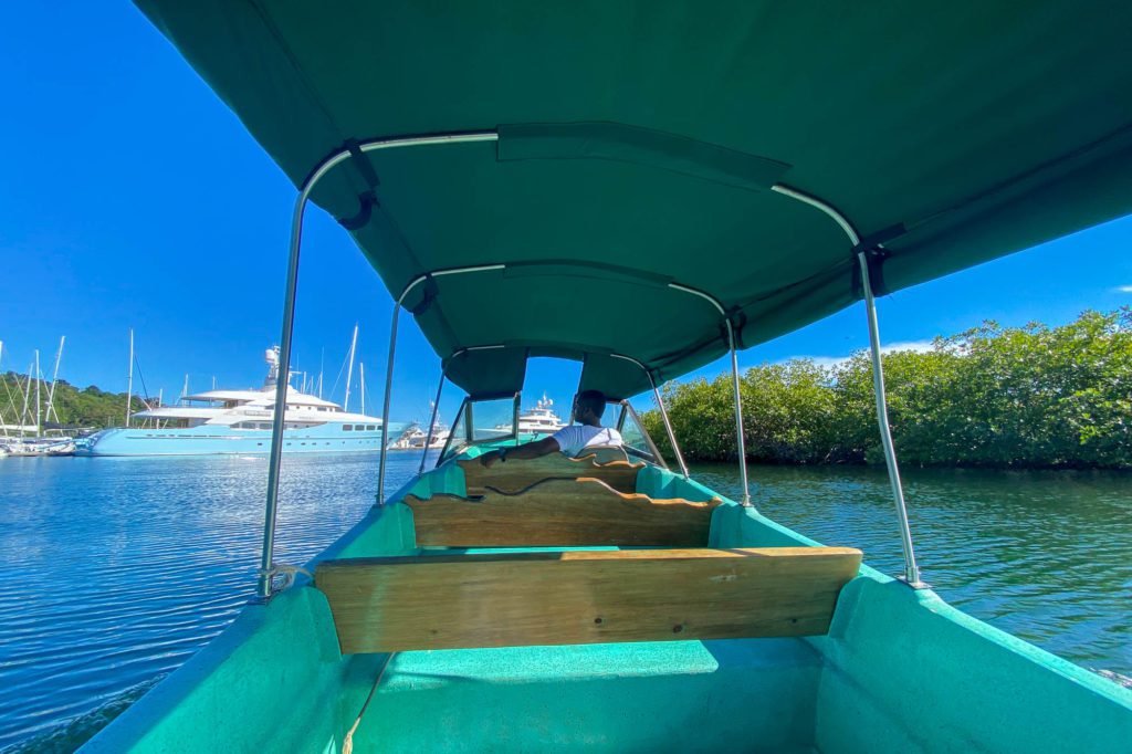 The image shows the interior of a green boat with a canopy, overlooking calm blue waters with yachts docked in the distance under a clear sky.