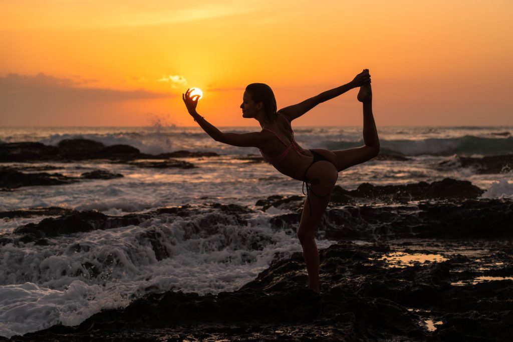 A person practices yoga on a rocky shore at sunset. They are balancing on one leg, with the other extended and held by the hand, silhouetted against the sun.