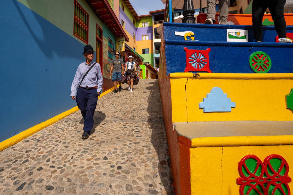 A person walks down a colorful street with vibrant buildings and a bright yellow staircase adorned with geometric patterns. Cobblestones pave the way under a clear sky.