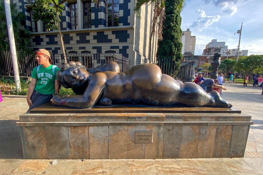 A bronze sculpture of a reclining figure lies in a public space with a person wearing green standing beside it, under a clear blue sky.
