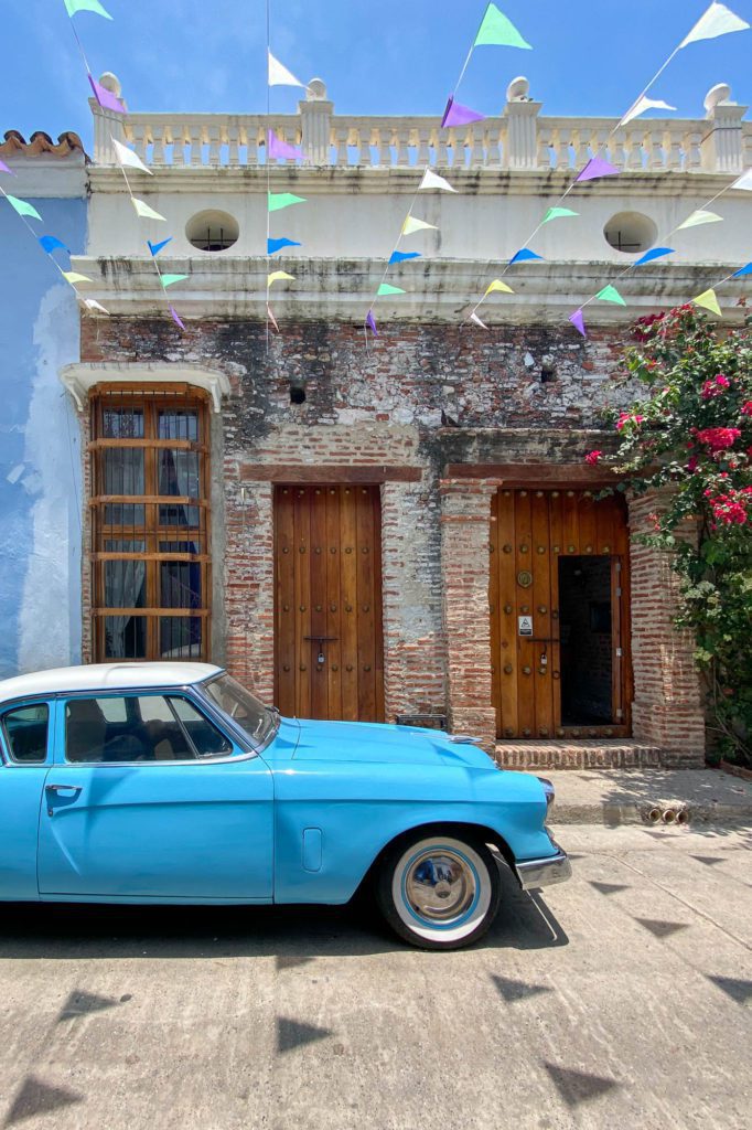 A vintage blue car is parked outside a weathered building with wooden doors, under colorful pennant flags, in a sunny, charming setting.