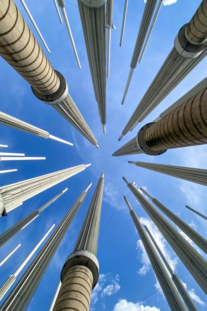 Upward view of numerous elongated vertical sculptures reaching towards a blue sky with white clouds, creating a dynamic perspective with converging lines.