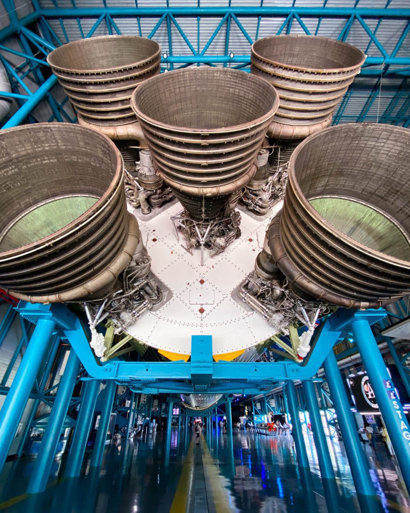 This is a close-up view of the massive rocket engines of a space vehicle, showcased in an exhibition with reflective floors and a blue steel structure.