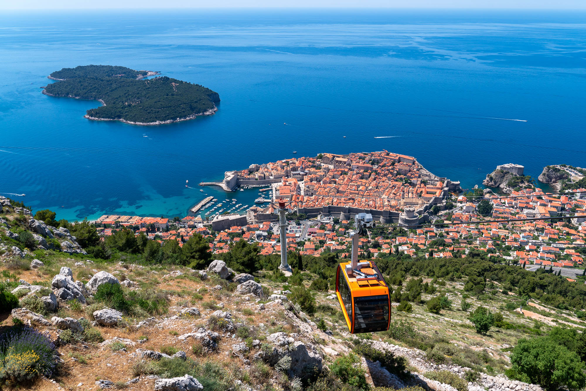 An aerial cable car descends towards a coastal city with orange roofs, surrounded by blue sea and a wooded island nearby, under a clear sky.