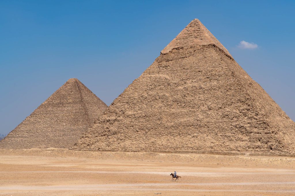 The image shows two of the Great Pyramids of Giza under a clear blue sky, with a person riding a horse in the vast desert foreground.